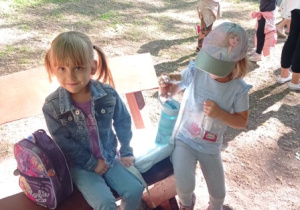 dwie dziewczynki , jedna siedzi na ławeczce, obok niej stoi druga