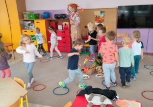 nauczycielka przebrana w jesienny stój prowadzi zabawę ruchową, obok są dzieci, na dywanie leżą obręcze