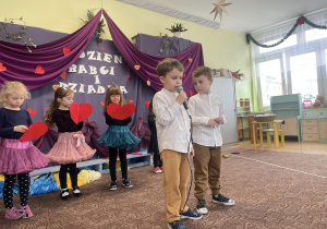 dwóch chłopców występuje przed zaproszonymi gośćmi, w tle dekoracja a także grupa dziewczynek, które trzymają w rączkach serca
