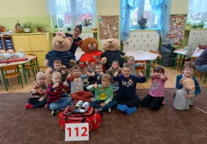 grupa dzieci, ratowniczka pozują do zdjęcia, obok misie, fantomy i nr alarmowy