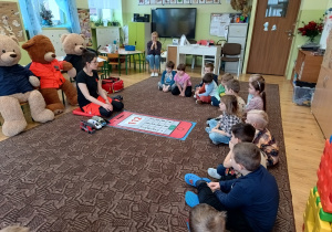 ratowniczka siedzi na dywanie, na podłodze leży plakat imitujący telefon, przed nią dzieci słuchają