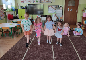 czworo dzieci trzyma się za ręce i idzie po dywanie, obok inne dzieci siedzą