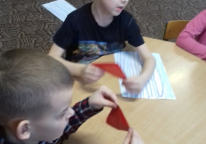 dwóch chłopców siedzi przy stoliku i zgina kolorowe papierowe origami -kwadraty