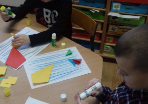 dwóch chłopców siedzi przy stoliku i przykleja papierowe origami -kwadraty