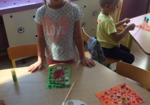 dziewczynka stoi przy stoliku, na którym leżą farby oraz praca plastyczna, którą wykonała