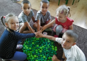 grupa dzieci siedzi na podłodze przy wykonanej z zielonych nakrętek "kropce"