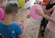 grupa dzieci stoi przy stoliku, trzymając w rękach balony, wykonują na papierze kropkowe stemple za ich pomocą