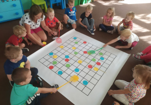 grupa dzieci siedzi na dywanie i układa kolorowe kółka na macie do kodowania. Obok jest nauczycielka