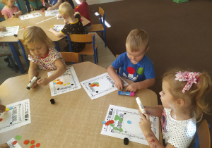dzieci siedzą przy stolikach i przyklejają kolorowe kółeczka na kartkach