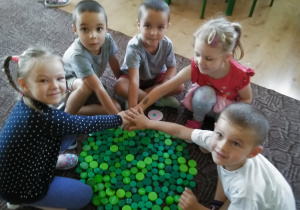 pięcioro dzieci siedzi na dywanie i prezentuje wykonaną kropkę z nakrętek