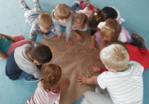 dzieci siedzą na dywanie przy grupowym plakacie i dokładają dłonie do odrysowanych szablonów