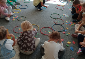 dzieci siedzą na dywanie i układają w kolorowych obręczach mozaikę geometryczną
