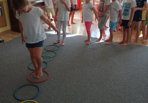grupa dzieci stoi na dywanie w strojach gimnastycznych, jedno dziecko przechodzi po torze z obręczy
