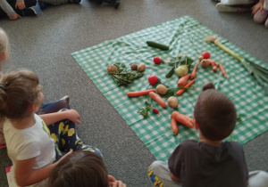 dzieci siedzą na dywanie , w środku na ceracie leżą warzywa