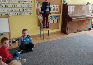 dzieci siedzą w kole, jedno dziecko stoi na krzesełku i recytuje wiersz
