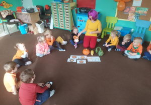 dzieci ubrane na pomarańczowo siedzą na dywanie i słuchają nauczycielki