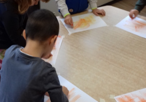 dzieci stoją przy stole i kreślą pomarańczową pastelą