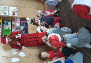 dzieci w mikołajowych czapkach siedzą na dywanie obok nich stoi ubrana na czerwono pani