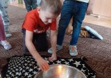 chłopiec kuca przy srebrnej misce z wodą, w środku jest zapalona świeca, dziecko wrzuca do wody złote grosiki