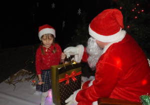w zimowej aranżacji siedzi Mikołaj na specjalnie przygotowanym fotelu, podaje dziecku prezent