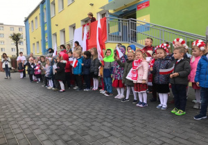 dzieci stoją przed budynkiem, przygotowują się do śpiewania hymnu