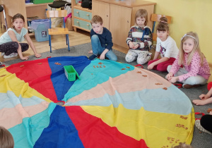dzieci siedzą na podłodze wokół kolorowej chusty, liczą na pestkach