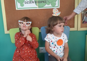 dwoje dzieci w ubraniach w kropki siedzi na krzesełkach, jedno z nich trzyma w rękach papierowe okulary