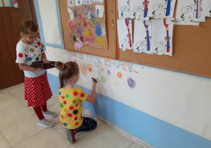 szatnia przedszkolna, długi papier przymocowany do ściany, dwie dziewczynki, jedna stoi i trzyma kredki, druga kuca i kreśli kropkę na papierze