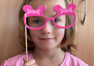 dziewczynka w różówej bluzeczce trzyma w rękach papierowe okulary i posuje do zdjęcia