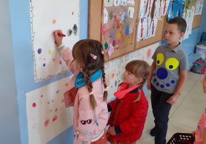 dzieci stoją w szatni, przy kartce przyklejonej do ściany, jedna z dziewczynek rysuje na papierze kropke