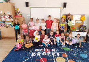 grupa dzieci stoi na dywanie w klasie, pozują do zdjęcia, w dłoniach trzymają balony w kropki, przed nimi na podłodze leży napis :"dzień kropki"