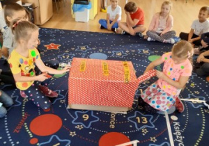 dzieci siedzą w kole na dywanie, na środku leży duże pudło ozdobione papierem w kropki, dwie dziewczynki otwierają je