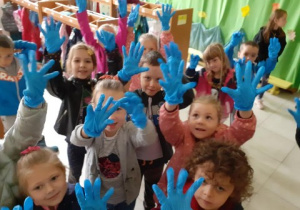 grupa dzieci w szatni przedszkolnej unosi do góry ręce, na dłoniach wszyscy mają założone gumowe rękawiczki