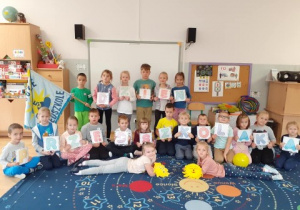 całą grupa dzieci pozuje do zdjęcia w klasie, trzymają w rękach litery, które układają się w napis "dzień przedszkolaka", niektóre mają w dłoniach balony
