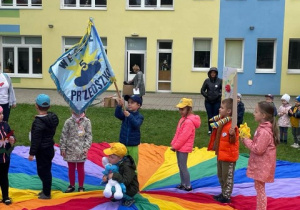 kilkoro dzieci stoi a środku chusty animacyjnej, trzymają w rękach emblematy i symbole grup, jeden z chłopców trzyma w rękach sztandar przedszkolny