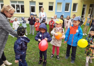 grupa dzieci na podwórku przedszkolnym, w dłoniach mają balony, nauczycielka podaje pinezkę do przebijania balonów, w środku są ukryte zadania