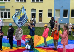podwórko przedszkolne, grupa dzieci stoi na chuście animacyjnej, chłopiec trzyma przedszkolny sztandar, reszta trzyma emblematy swoich grup przedszkolnych