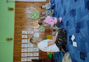 biblioteka, dzieci siedzą na dywanie, przy bibliotekarce siedzi dziewczynka, która losuje coś z worka
