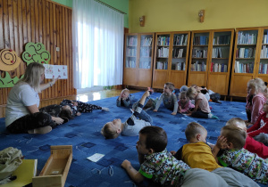 biblioteka, dzieci leżą na dywanie , obok pani bibliotekarka pokazuje ilustracje w ksiażce