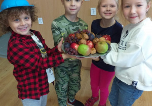 czworo dzieci stoi, trzymają w rękach tacę z owocami