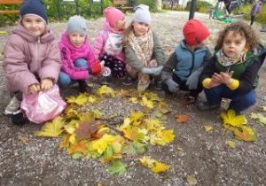 grupa dzieci kuca, przed nimi leżą zebrane przez nie żółte liście