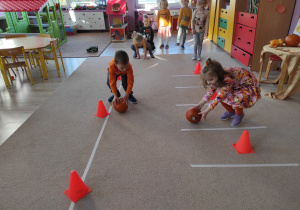 dzieci biorą udział w zabawach sportowych, dwoje z nich toczy dynie po dywanie