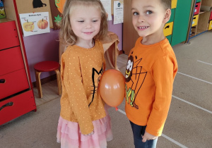 dwoje dzieci trzyma między brzuszkami pomarańczowy balon