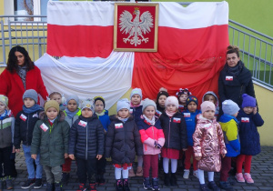 Dzieci z grupy Biedronki i ich panie, stoją przed wejściem do przedszkola. Na poręczy przy wejściu powieszone są biało-czerwone flaki oraz godło