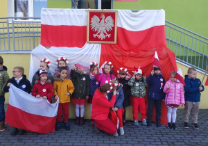 Dzieci z grupy Słoneczka i ich nauczycielka, stoją przed wejściem do przedszkola. Na poręczy przy wejściu powieszone są biało-czerwone flaki oraz godło
