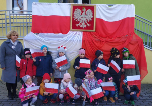 Dzieci z grupy Duszki i ich nauczycielka, stoją przed wejściem do przedszkola. Na poręczy przy wejściu powieszone są biało-czerwone flaki oraz godło