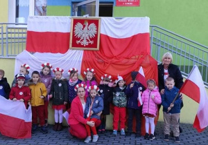 Dzieci z grupy Słoneczka, ich nauczycielka i pani dyrektor, stoją przed wejściem do przedszkola. Na poręczy przy wejściu powieszone są biało-czerwone flaki oraz godło
