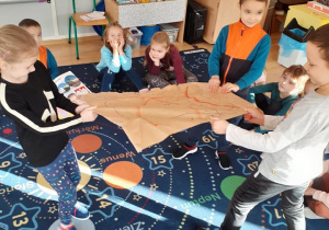 grupa dzieci siedzi na dywanie, troje z nich rozrywa papier, na którym narysowany jest kontur Polski