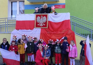 grupa dzieci stoi przed wejsciem do przedszkola, chłopcy trzymają flagę, dziewczynki mają na głowach biało-czerwone wianki