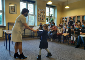 dziewczynka odbiera nagrodę od pracownika biblioteki, w tle siedzą inni uczestnicy konkursu z opiekunami